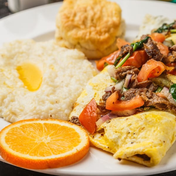 A breakfast plate with a loaded omelet, grits with butter, a biscuit, and an orange slice.