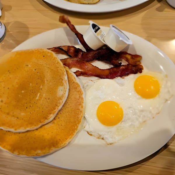 A breakfast plate with two pancakes, crispy bacon, two sunny-side-up eggs, and creamer cups.