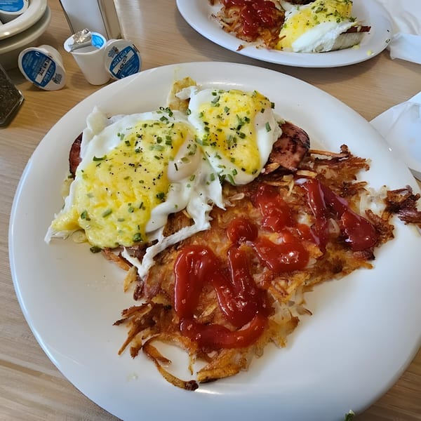 Two plates of breakfast hash browns, eggs Benedict, and sausage, with ketchup on the hash browns.