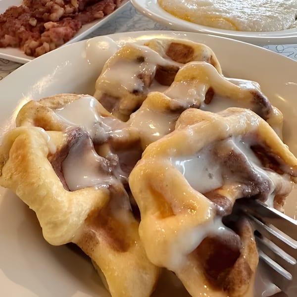 Cinnamon roll waffles with white icing on a plate, with a fork and other breakfast items.