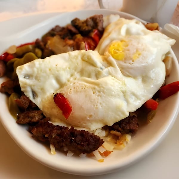 A breakfast skillet with two sunny-side-up eggs, sausage, hash browns, and peppers in a white bowl.