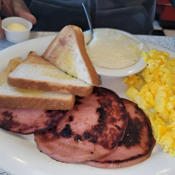 A breakfast plate with grilled ham, scrambled eggs, grits, and buttered toast from That Flippin' Egg.
