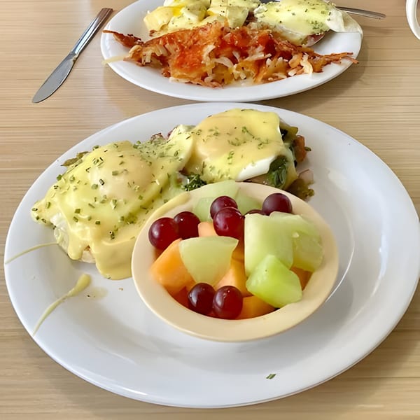 Two plates of breakfast, one with Eggs Benedict and fruit, the other with hash browns.