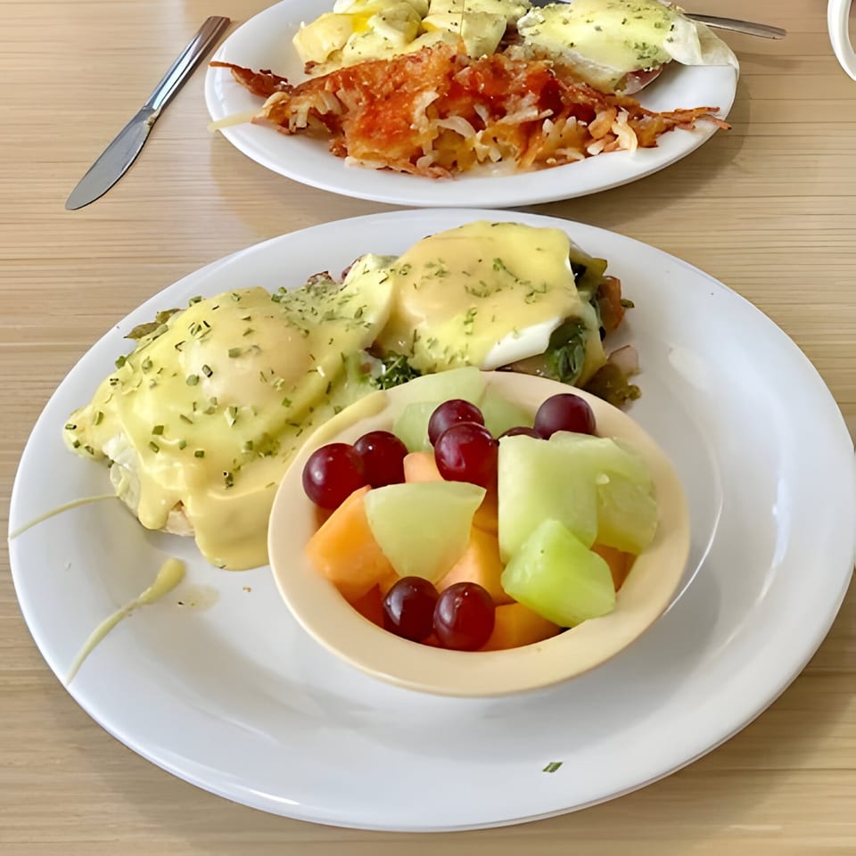 Two plates of breakfast, one with Eggs Benedict and fruit, the other with hash browns.