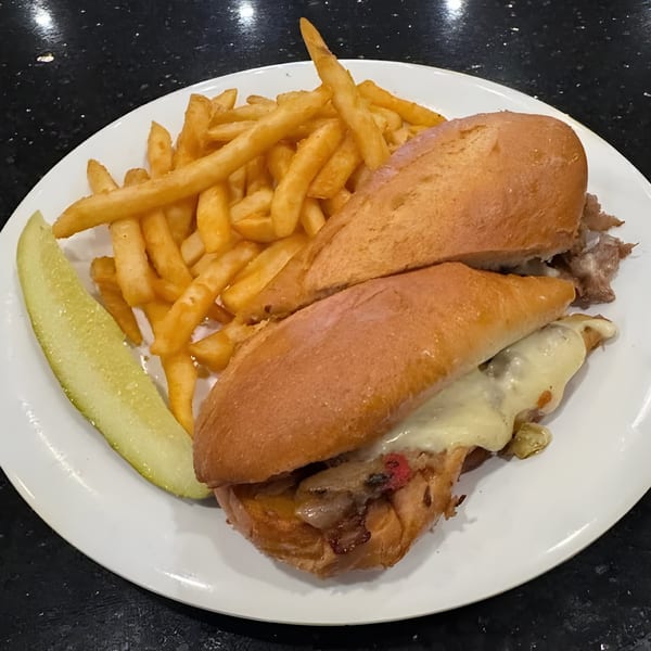 Cheesesteak sandwich with melted cheese, fries, and a pickle spear on a white plate.