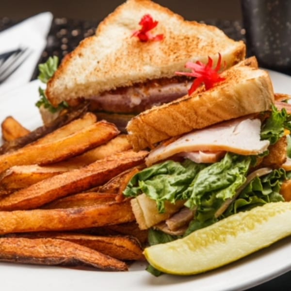 Turkey club sandwich with lettuce, tomato, and bacon, served with a side of fries and a pickle.
